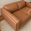 Augo vegan suede two-seater loveseat sofa upholstered in warm tan leather, shown in a styled interior setting with a textured cream rug and light wood flooring.