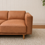 A close-up of a tan Hugo Vegan Suede loveseat sofa with wooden legs, set on a textured cream rug against a white paneled wall.