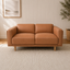 Hugo vegan suede loveseat sofa in warm tan leather with a light wood frame and legs, positioned on a textured cream rug in a bright, modern room with white paneled walls.