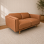 A mid-century modern loveseat sofa upholstered in warm camel brown leather, set on a textured cream rug against a white paneled wall.