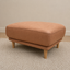 Hugo vegan suede ottoman in a warm tan or camel fabric, supported by four tapered light wood legs, set on a textured light beige rug against a neutral beige wall.