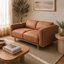 A two-seater loveseat sofa upholstered in warm tan faux leather, styled in a bright, modern living room with a wooden coffee table, woven rug, and potted plants.