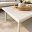 A modern low-profile srectangle outdoor coffee table in a warm off-white finish, styled with a wooden tray and potted plant on top, set in a bright, sunlit living room.